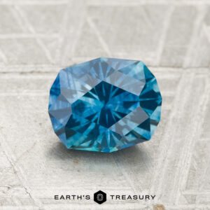 A 1.46-carat heated Montana sapphire, round and faceted, sits on a textured gray surface with "EARTH'S TREASURY" displayed at the bottom.