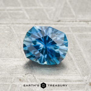A 1.17-carat heated Montana sapphire is showcased on a textured gray surface, with the "Earth's Treasury" logo and text displayed at the bottom.