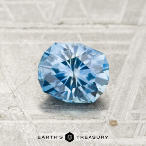 A faceted, round blue 1.05-carat Montana Sapphire (heated) rests on a textured metallic surface. "EARTH'S TREASURY" appears at the bottom of the image.