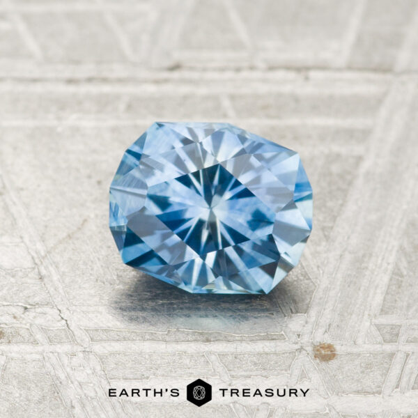 1.05-Carat Montana Sapphire (Heated) A faceted, round blue 1.05-carat Montana Sapphire (heated) rests on a textured metallic surface. "EARTH'S TREASURY" appears at the bottom of the image.