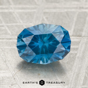 A 2.29-carat heated Montana sapphire is displayed on a textured gray surface, with "EARTH'S TREASURY" and a logo shown at the bottom.