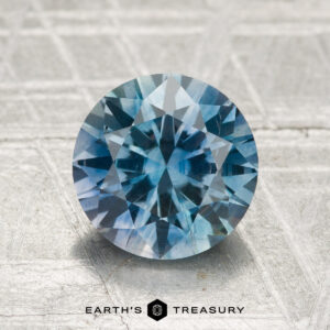 A 2.26-carat heated Montana sapphire, cut in a round brilliant style, sits on a textured light gray surface with "EARTH'S TREASURY" displayed at the bottom.