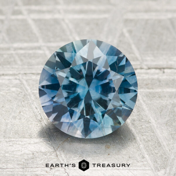 2.26-Carat Montana Sapphire (Heated) A 2.26-carat heated Montana sapphire, cut in a round brilliant style, sits on a textured light gray surface with "EARTH'S TREASURY" displayed at the bottom.