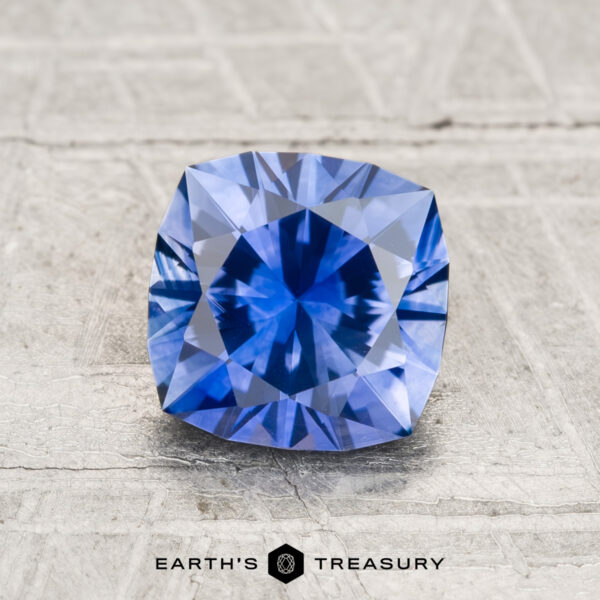 1.63-Carat Montana Sapphire (Heated) A 1.63-carat heated Montana sapphire, cushion-cut and blue, rests on a textured light gray surface with "EARTH'S TREASURY" shown below.