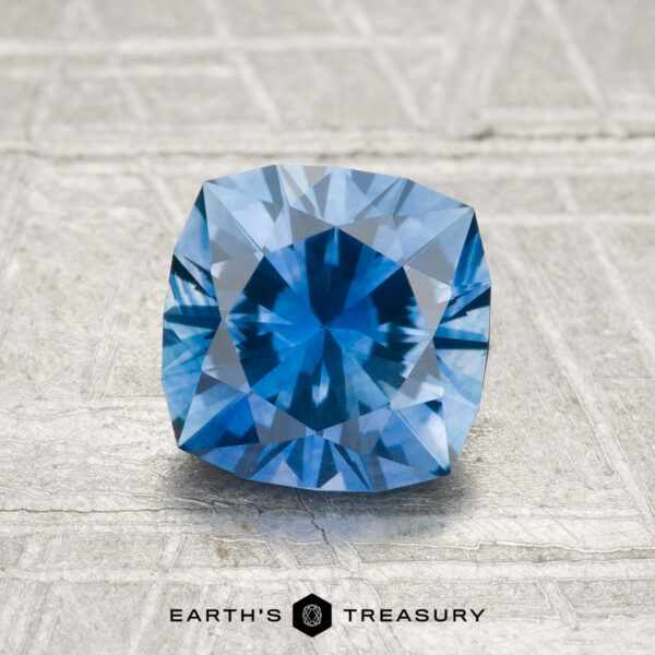 1.63-Carat Montana Sapphire (Heated) A 1.63-carat heated Montana sapphire with a cushion cut is displayed on a textured gray surface, featuring the "Earth's Treasury" logo and text at the bottom.