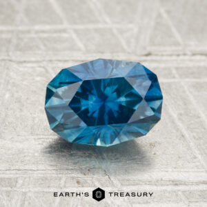A 1.76-carat heated Montana sapphire is displayed on a textured surface with the "Earth's Treasury" logo beneath it.