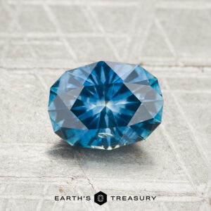 A faceted oval blue 1.46-carat heated Montana sapphire with a starburst pattern is displayed on a textured gray surface, with the "EARTH'S TREASURY" logo below the stone.