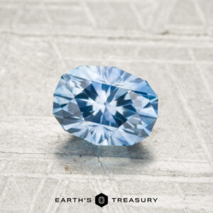 A 1.02-carat heated Montana sapphire, cut in a faceted oval shape, is displayed on a textured light surface with "EARTH'S TREASURY" beneath it.