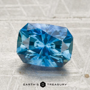 A 2.60-carat heated Montana sapphire in a blue, rectangular cut with intricate facets rests on a textured, light surface. The "Earth's Treasury" logo is visible at the bottom.