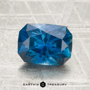 A 2.33-carat heated Montana sapphire in a rectangular cut with multiple facets sits on a textured gray surface. "Earth's Treasury" is shown at the bottom.