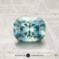 A 1.34-carat heated Montana sapphire with blue-green hues and a rectangular step cut is displayed on a textured light gray surface above the Earth's Treasury logo.