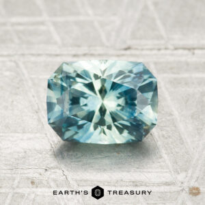 A 1.34-carat heated Montana sapphire with blue-green hues and a rectangular step cut is displayed on a textured light gray surface above the Earth's Treasury logo.