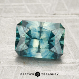 A 2.51-carat heated Montana sapphire with geometric facets is displayed on a textured gray surface, with the Earth’s Treasury logo at the bottom.