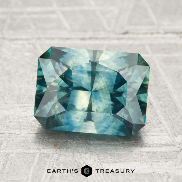 2.51-Carat Montana Sapphire (Heated) A 2.51-carat heated Montana sapphire with geometric facets is displayed on a textured gray surface, with the Earth’s Treasury logo at the bottom.
