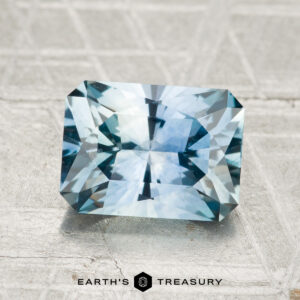 A 2.32-carat rectangular, faceted Montana sapphire (heated) with a vivid blue hue is displayed on a textured gray surface, featuring the text "EARTH'S TREASURY" underneath.