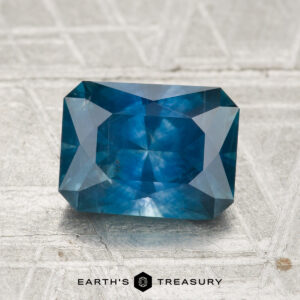 A rectangular, faceted 2.17-carat heated Montana sapphire with a blue hue is displayed on a textured light surface, featuring the "EARTH'S TREASURY" logo beneath.