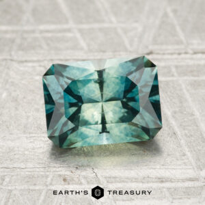 A faceted, rectangular blue-green 1.98-carat heated Montana Sapphire rests on a textured gray surface, labeled "EARTH'S TREASURY" at the bottom.