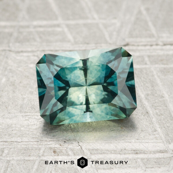 1.98-Carat Montana Sapphire (Heated) A faceted, rectangular blue-green 1.98-carat heated Montana Sapphire rests on a textured gray surface, labeled "EARTH'S TREASURY" at the bottom.