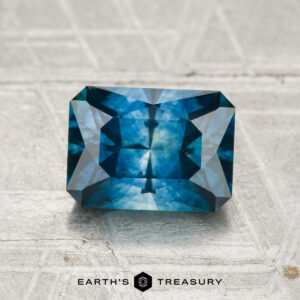A 1.88-carat heated Montana sapphire, cut in a rectangular faceted shape, rests on a textured metallic surface with the Earth’s Treasury logo below.