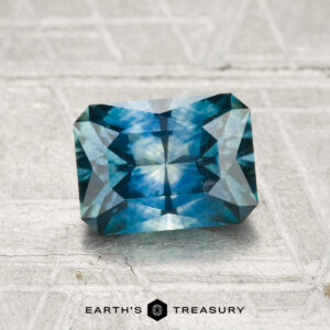 A 1.54-carat heated Montana sapphire with a rectangular brilliant cut sits on a textured light gray surface, "EARTH'S TREASURY" displayed below.