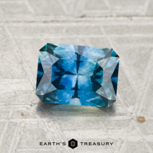 A rectangular, faceted blue 1.49-carat Montana Sapphire (Heated) rests on a textured surface featuring the "EARTH'S TREASURY" text and logo at the bottom.