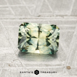 A 1.47-carat heated Montana sapphire with green and yellow hues rests on a textured silver surface; the "Earth's Treasury" logo appears at the bottom.