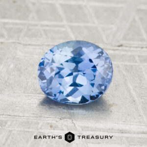 A 1.35-carat Ceylon sapphire, faceted in an oval shape, rests on a textured gray surface viewed from above. "EARTH'S TREASURY" appears at the bottom of the image.