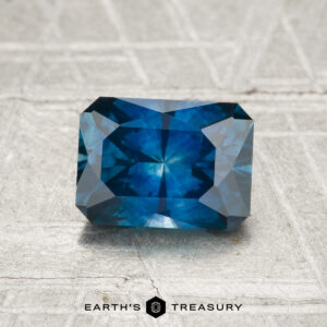 A 1.42-carat heated Montana sapphire with a rectangular, faceted cut is displayed on a textured, light background, featuring the text "EARTH'S TREASURY" at the bottom.