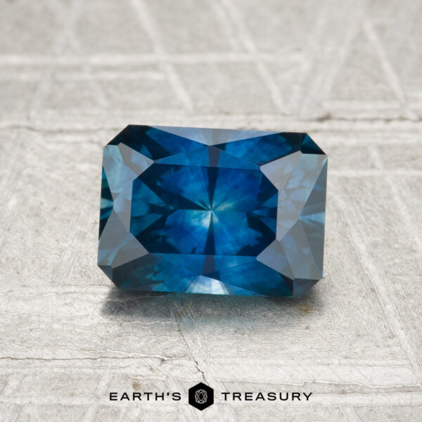1.42-Carat Montana Sapphire (Heated) A 1.42-carat heated Montana sapphire with a rectangular, faceted cut is displayed on a textured, light background, featuring the text "EARTH'S TREASURY" at the bottom.