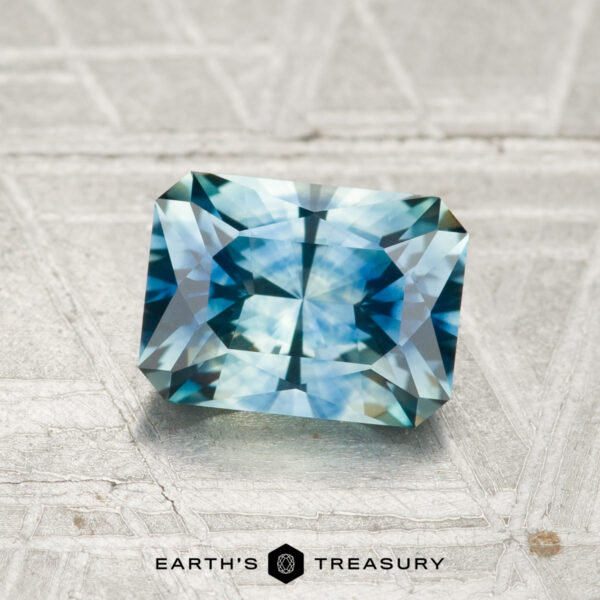1.39-Carat Montana Sapphire (Heated) A 1.39-carat heated Montana sapphire with radiant facets and a blue-green hue rests on a textured, light surface. The "EARTH'S TREASURY" logo and text are visible at the bottom.