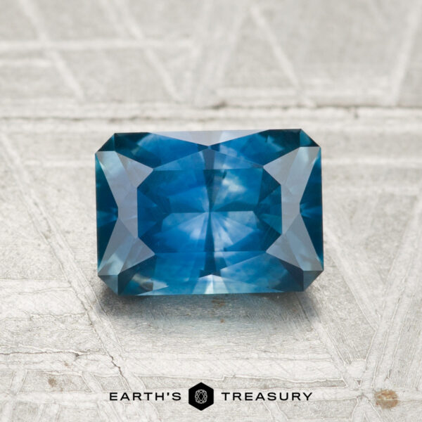 A 1.37-carat heated Montana sapphire, rectangular and blue with facet cuts, rests on a textured light surface. "Earth's Treasury" appears at the bottom of the image.