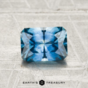 A 1.35-carat rectangular, faceted Montana Sapphire (heated) is displayed on a light textured surface with the text "EARTH'S TREASURY" below it.