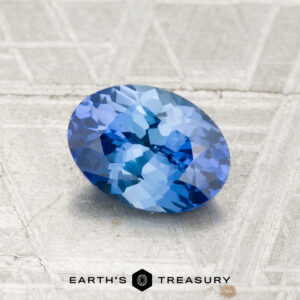 A 1.17-carat faceted oval Ceylon sapphire gemstone resting on a textured gray surface, with the “EARTH’S TREASURY” text and logo displayed at the bottom.