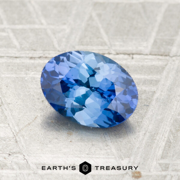 1.17-Carat Ceylon Sapphire A 1.17-carat faceted oval Ceylon sapphire gemstone resting on a textured gray surface, with the “EARTH’S TREASURY” text and logo displayed at the bottom.
