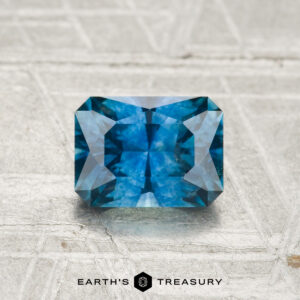 A 1.22-carat heated Montana sapphire, cut in a rectangular shape with multiple facets, rests on a textured light surface with "EARTH'S TREASURY" written below.
