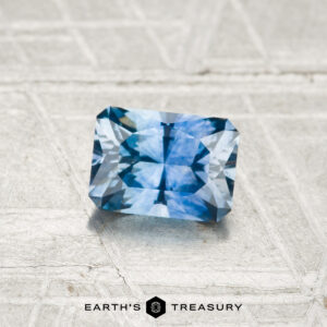A 0.76-carat heated Montana sapphire with radiant facets is displayed on a textured light surface, featuring the text "EARTH'S TREASURY" and a small hexagonal logo below.