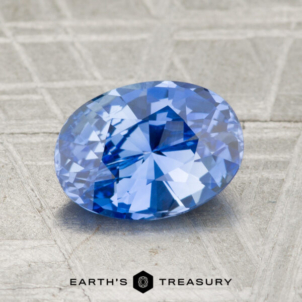 A 1.72-carat Ceylon Sapphire, oval-cut, rests on a textured gray surface with "EARTH'S TREASURY" and a hexagonal logo at the bottom.