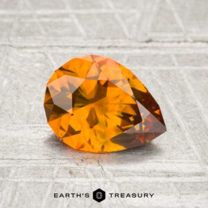 A 1.37-carat heated Montana sapphire, faceted in a pear shape with an orange hue, is displayed on a textured gray surface with the text "Earth's Treasury" below.