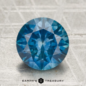 A round, faceted 3.35-carat Montana Sapphire (heated) with a vivid blue hue is showcased on a textured surface, featuring the Earth's Treasury logo at the bottom.