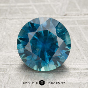 A 2.92-carat heated Montana sapphire in round brilliant cut and blue-green hue is displayed on a textured surface, with "EARTH'S TREASURY" text featured at the bottom.