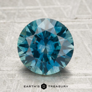 A 2.80-carat heated Montana sapphire, round brilliant-cut in blue-green, is displayed on a textured light surface with "EARTH'S TREASURY" at the bottom.
