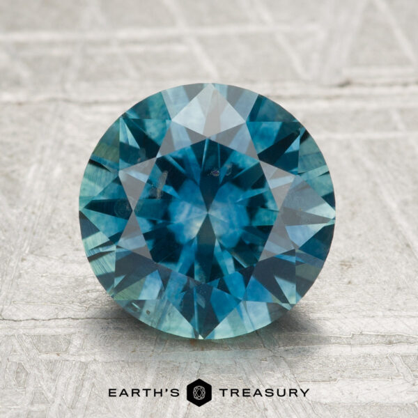 2.80-Carat Montana Sapphire (Heated) A 2.80-carat heated Montana sapphire, round brilliant-cut in blue-green, is displayed on a textured light surface with "EARTH'S TREASURY" at the bottom.
