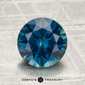 A round, faceted 2.60-carat heated Montana sapphire is displayed on a textured metallic surface with "Earth's Treasury" below.
