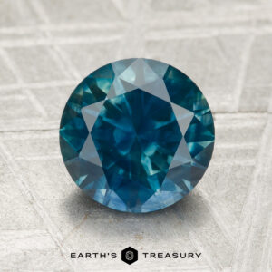 A round, faceted 2.42-carat heated Montana sapphire with a blue-green hue is displayed on a textured gray surface, featuring the "EARTH'S TREASURY" logo and text at the bottom.