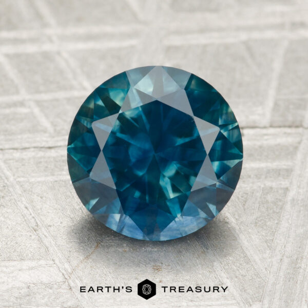 2.42-Carat Montana Sapphire (Heated) A round, faceted 2.42-carat heated Montana sapphire with a blue-green hue is displayed on a textured gray surface, featuring the "EARTH'S TREASURY" logo and text at the bottom.