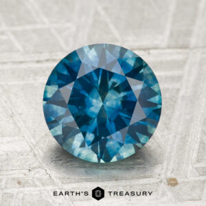 A 2.41-carat heated Montana Sapphire, round brilliant-cut and blue in color, displayed on a textured surface with the "Earth's Treasury" logo and text at the bottom.