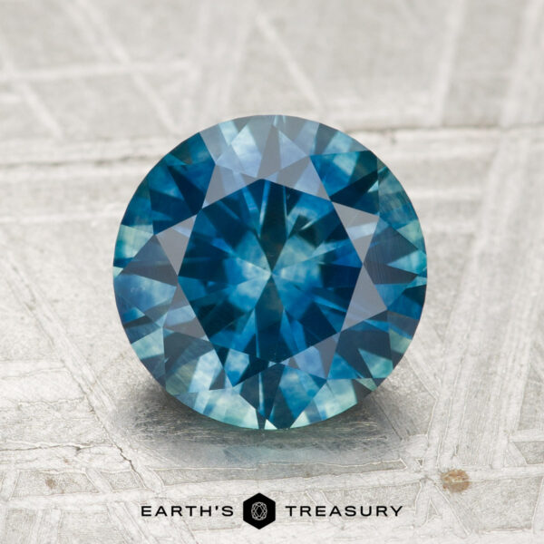 2.41-Carat Montana Sapphire (Heated) A 2.41-carat heated Montana Sapphire, round brilliant-cut and blue in color, displayed on a textured surface with the "Earth's Treasury" logo and text at the bottom.