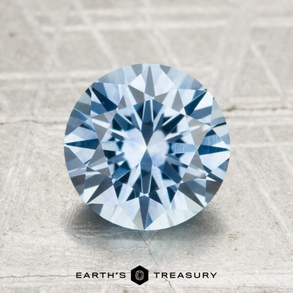 2.40-Carat Montana Sapphire (Heated) A 2.40-carat heated Montana sapphire in round brilliant cut is shown on a textured light surface with the Earth’s Treasury logo below.