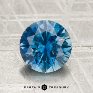 A 2.39-carat heated Montana sapphire, cut in a round brilliant style, is displayed on a textured light surface with "Earth's Treasury" below it.