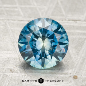 A 2.33-carat heated Montana sapphire, round-cut and blue, is displayed on textured gray with the "EARTH'S TREASURY" text and logo shown below.